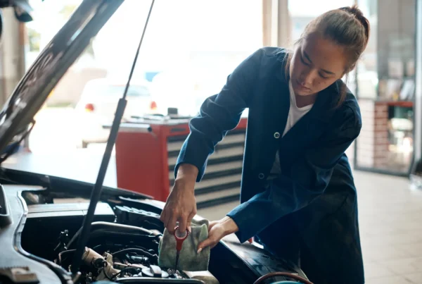 A female mechanic in a blue work coat checks a car's oil dipstick with a rag under the raised hood in an auto shop.