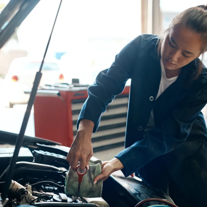 A female mechanic in a blue work coat checks a car's oil dipstick with a rag under the raised hood in an auto shop.