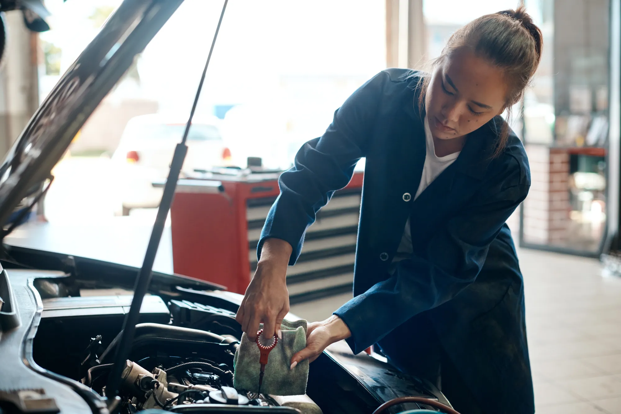 A female mechanic in a blue work coat checks a car's oil dipstick with a rag under the raised hood in an auto shop.